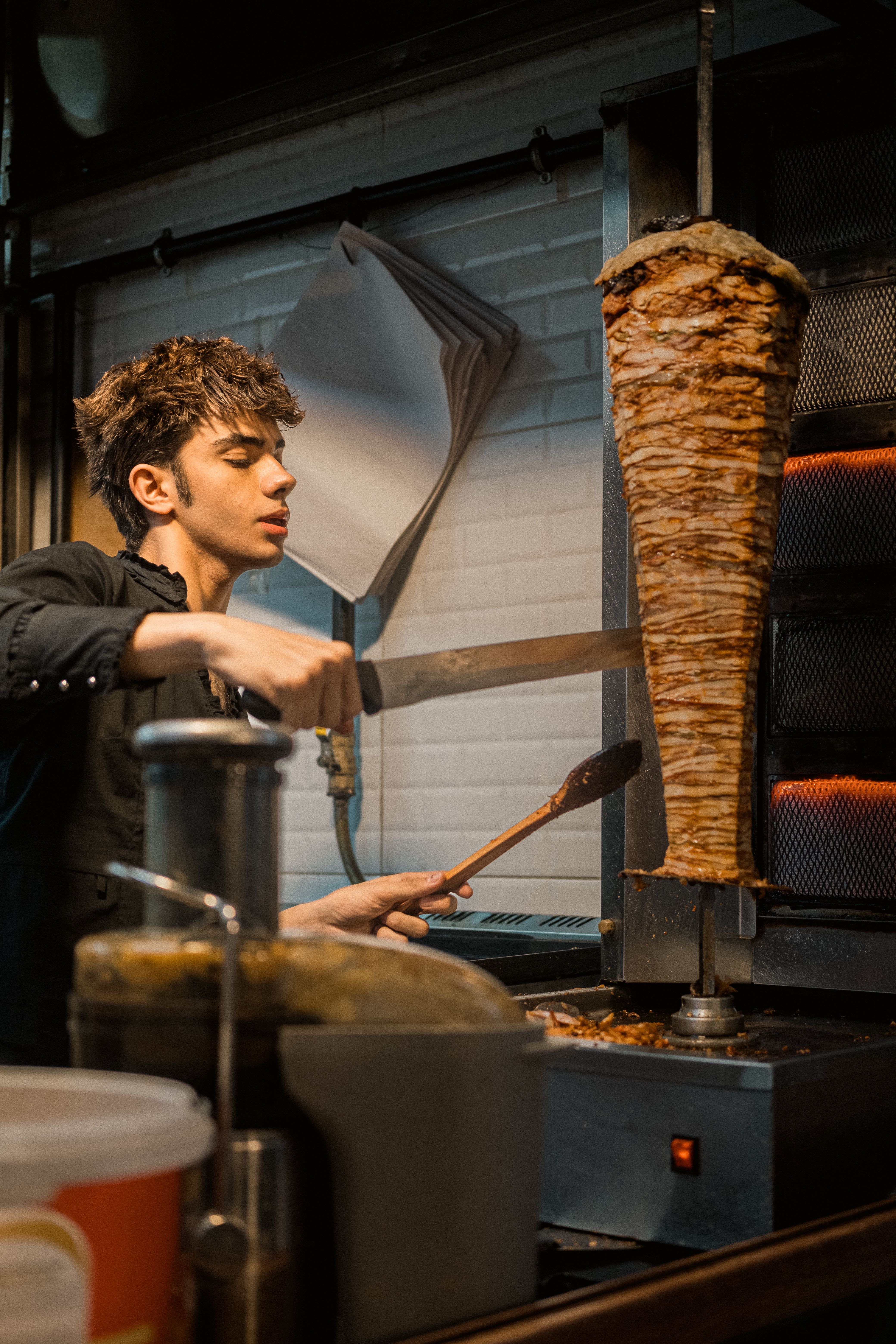 Young man slicing meat from a vertical rotisserie spit in a restaurant kitchen.